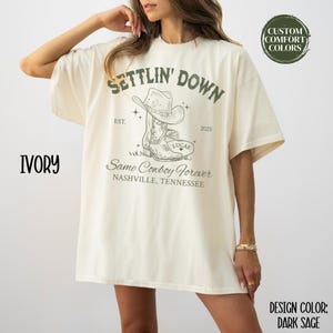 May include: Ivory oversized t-shirt with a graphic design of a cowboy boot and hat. The text on the shirt reads "Settlin' Down Est. 2025 Same Cowboy Forever Nashville, Tennessee".