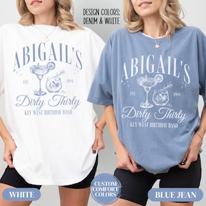Pode incluir: Duas mulheres vestindo camisetas com o texto "Abigail's Dirty Thirty Key West Birthday Bash" impresso nelas. As camisetas s&atilde;o de cor branca e azul jeans. O texto est&aacute; em uma fonte de estilo vintage com um desenho de uma ta&ccedil;a de margarita e uma ta&ccedil;a de coquetel.