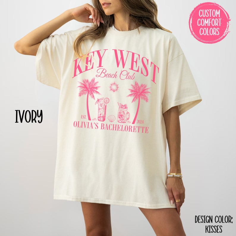 Pu&ograve; includere: Una maglietta bianca con un design grafico rosa che dice "Key West Beach Club Est. 2021 Olivia's Bachelorette". La grafica include anche palme, un sole e due cocktail.