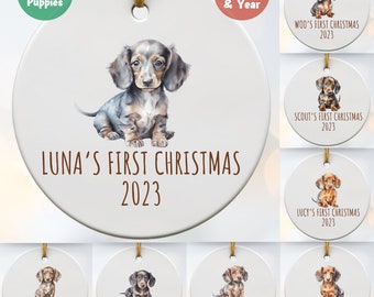 May include: Twelve dachshund puppies, each with a personalized Christmas ornament featuring a watercolor illustration of a dachshund puppy. The ornaments are white with brown text that reads "[puppy's name]'s First Christmas 2023".