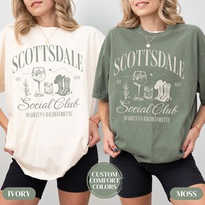 May include: Two t-shirts in ivory and moss green with a graphic design of a cactus, a glass of wine, a shot glass, and cowboy boots. The text on the shirts reads "Scottsdale Social Club Est. 2024 Madelyn's Bachelorette".