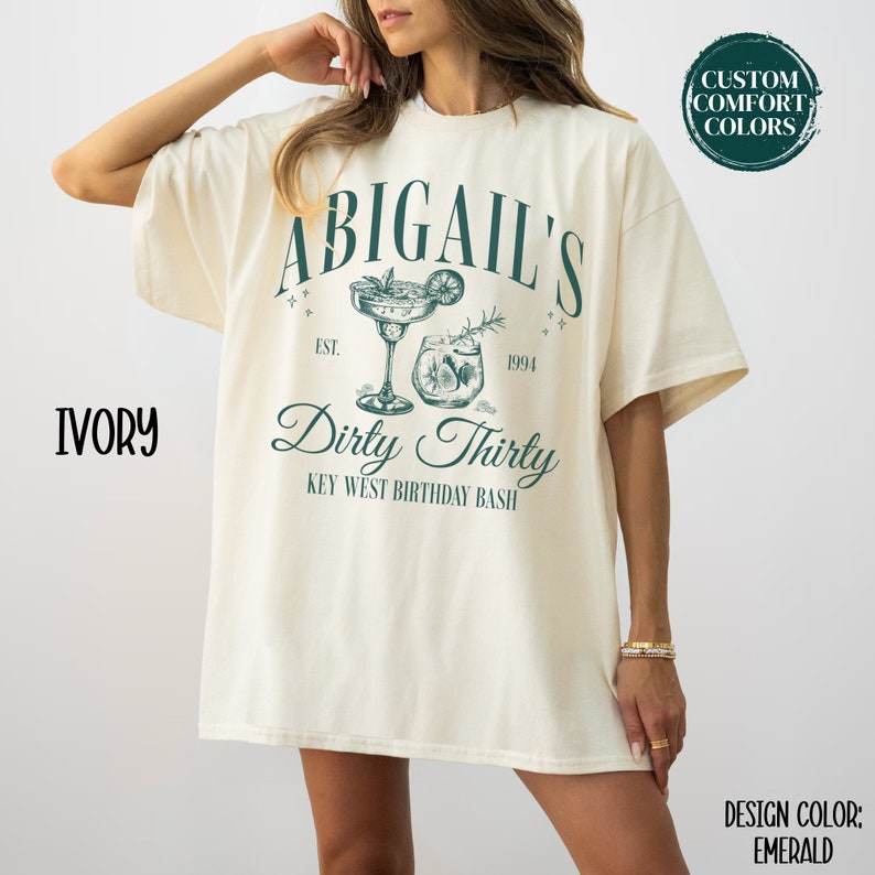 Pode incluir: Camiseta de cor marfim com um design verde que diz "Abigail's Dirty Thirty Key West Birthday Bash" com um desenho de dois coquet&eacute;is.