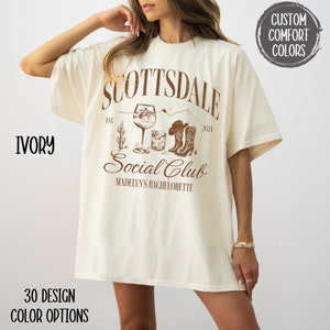 May include: Ivory oversized t-shirt with a brown graphic design that says "Scottsdale Social Club" with a cactus, cowboy boots, and a cocktail glass. The text also says "Est. 2024" and "Madelyn's Bachelorette".