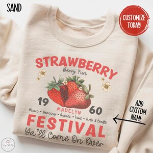 May include: A beige sweatshirt with a graphic design of strawberries and the text "STRAWBERRY Berry Fun 19 MADELYN 60 FESTIVAL Ya'll Come On Over" in red and black. The text "ADD CUSTOM NAME" is also included on the sweatshirt.