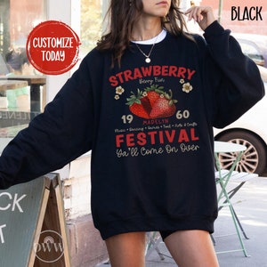 May include: A black sweatshirt with a graphic design featuring a strawberry festival. The design includes the text "Strawberry Berry Fun", "Madelyn", "19", "60", "Music Dancing Games Food Arts & Crafts", "Festival", and "Ya'll Come On Over".