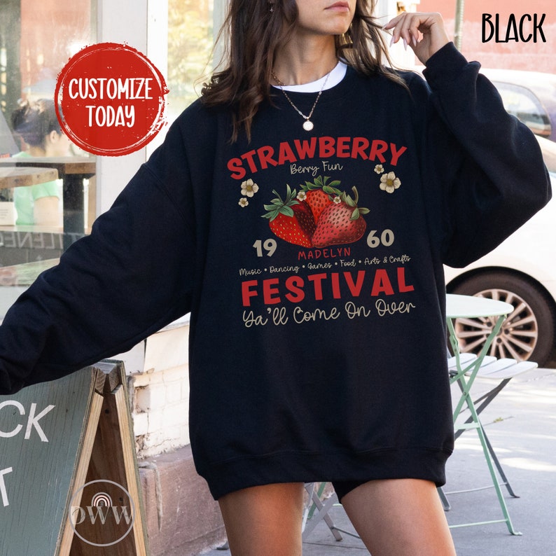May include: A black sweatshirt with a graphic design featuring a strawberry festival. The design includes the text "Strawberry Berry Fun", "Madelyn", "19", "60", "Music Dancing Games Food Arts & Crafts", "Festival", and "Ya'll Come On Over".
