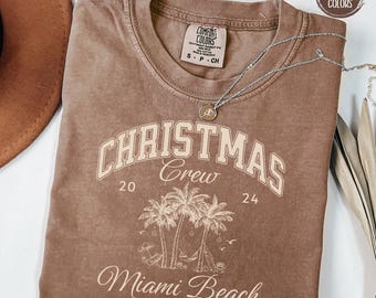 May include: Espresso brown t-shirt with a vintage-style graphic design that says "Christmas Crew 20 24 Miami Beach Florida" in ivory lettering. The design features palm trees and a hammock.