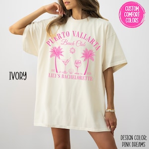 Pu&ograve; includere: Una maglietta bianca con un design grafico rosa che dice "Puerto Vallarta Beach Club" con palme, bevande e il testo "Est. 2024 Lily's Bachelorette".