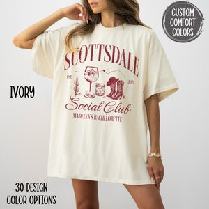 May include: A white t-shirt with a red graphic design that says "Scottsdale Social Club" with a wine glass, cactus, and cowboy boots. The text "Est. 2024" and "Madelyn's Bachelorette" are also included in the design.