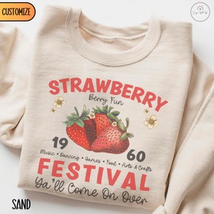May include: A beige sweatshirt featuring a red and black graphic design that reads "Strawberry Berry Fun 19 60 Festival Ya'll Come On Over". The graphic also includes a cluster of red strawberries with green leaves and white flowers.