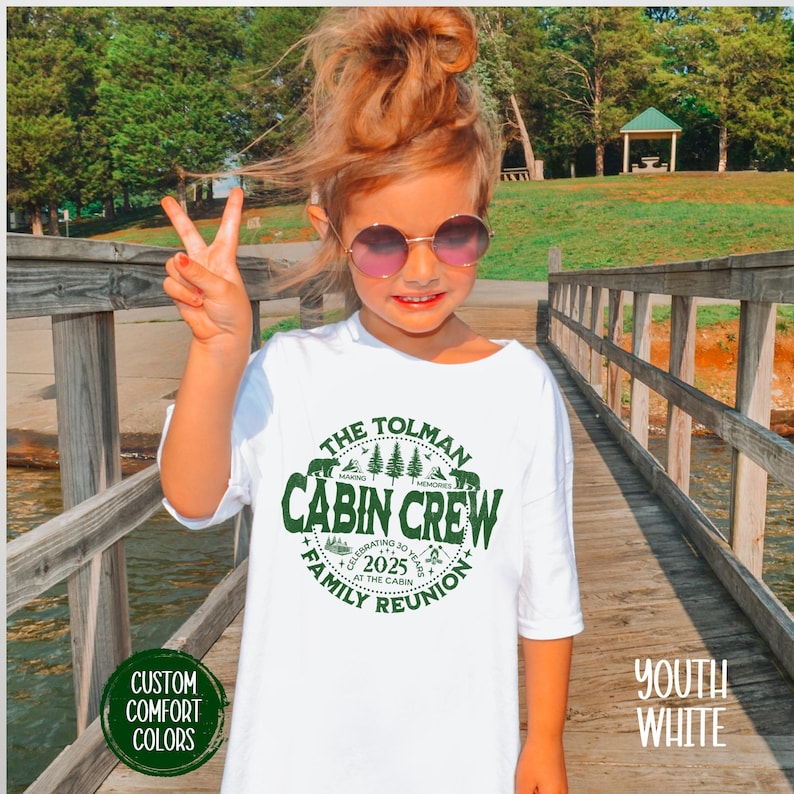 May include: A white t-shirt with a green circular graphic that reads "The Tolman Cabin Crew Family Reunion 2025". The shirt is labeled "Youth White" and "Custom Comfort Colors". A young person is wearing the shirt and giving a peace sign.