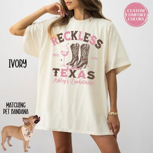 May include: Ivory-colored t-shirt with a graphic design featuring the word "Reckless" above a pair of cowboy boots and a wine glass. The text "Texas" and "Ashley's Bachelorette" are also included. A matching pet bandana is also shown.
