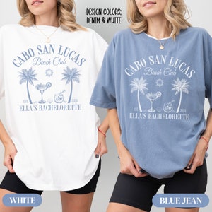 Pu&ograve; includere: Due donne indossano magliette oversize con un design grafico blu che dice "Cabo San Lucas Beach Club" con palme, un bicchiere di margarita e il testo "Est. 2024 Ella's Bachelorette". La maglietta a sinistra &egrave; bianca e la maglietta a destra &egrave; blu jeans.