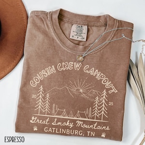 May include: Espresso-colored t-shirt with the text "COUSIN CREW CAMPOUT" and a graphic of mountains, trees, and tents. The text "Great Smoky Mountains GATLINBURG, TN" is also visible. A silver necklace with a pendant is draped across the shirt.