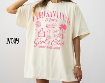 May include: Ivory t-shirt with a pink graphic design that says "Cabo San Lucas + Mexico" with a margarita, palm tree, and the text "Girl's Club Maddie's Bachelorette".