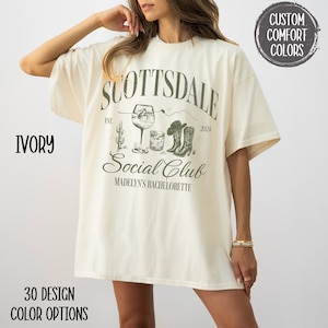 May include: Ivory oversized t-shirt with a graphic design featuring the text "Scottsdale Social Club" with a cactus, a glass of wine, a cocktail glass, and cowboy boots. The text "Est. 2024" and "Madelyn's Bachelorette" are also included in the design.