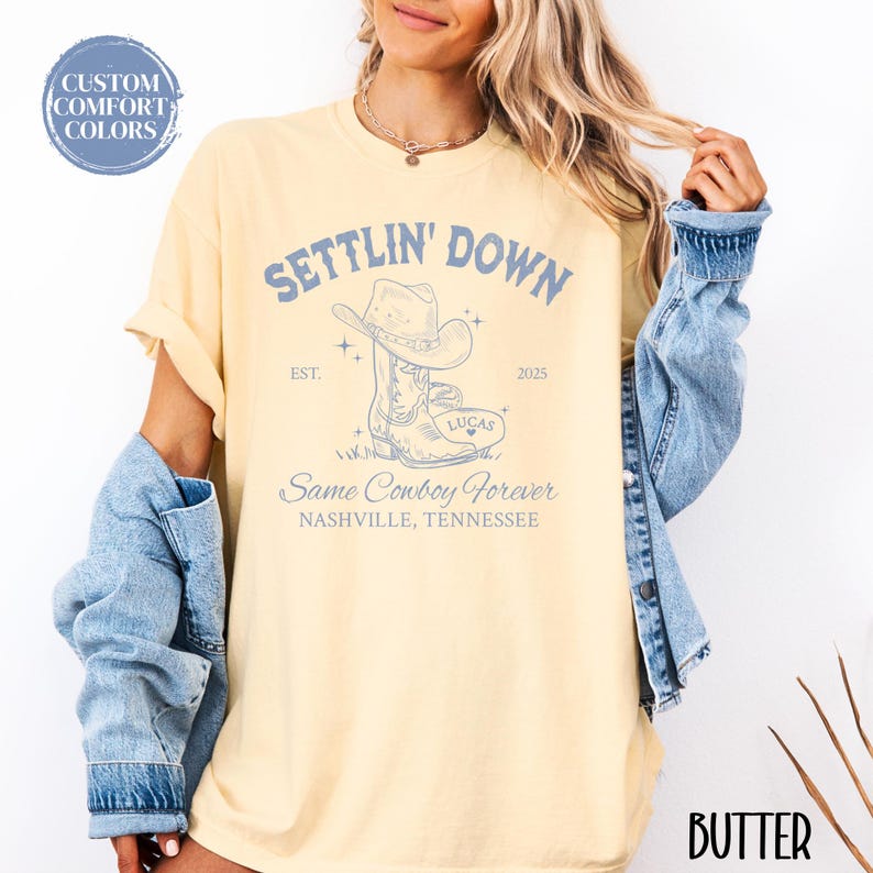 May include: A yellow t-shirt with the text "Settlin' Down" and "Same Cowboy Forever" in blue. The shirt also features a drawing of a cowboy boot with a cowboy hat on top of it. The text "EST. 2025" and "Nashville, Tennessee" are also on the shirt.
