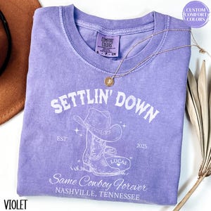May include: A purple t-shirt with a white graphic of a cowboy boot and hat. The text "Settlin' Down" is above the graphic, and the text "Same Cowboy Forever" is below the graphic. The text "EST. 2025" is on the right side of the graphic. The text "Lucas" is on the boot.