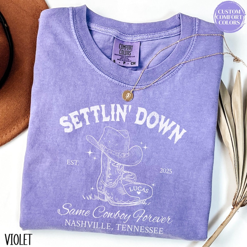 May include: A purple t-shirt with a white graphic of a cowboy boot and hat. The text "Settlin' Down" is above the graphic, and the text "Same Cowboy Forever" is below the graphic. The text "EST. 2025" is on the right side of the graphic. The text "Lucas" is on the boot.