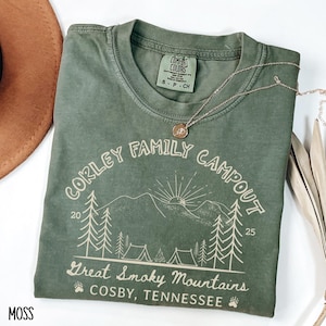 May include: A moss green t-shirt with the text "CORLEY FAMILY CAMPOUT" and "Great Smoky Mountains, Cosby, Tennessee" printed on it. The design includes trees, mountains, and tents. A silver necklace with a pendant is on the shirt.