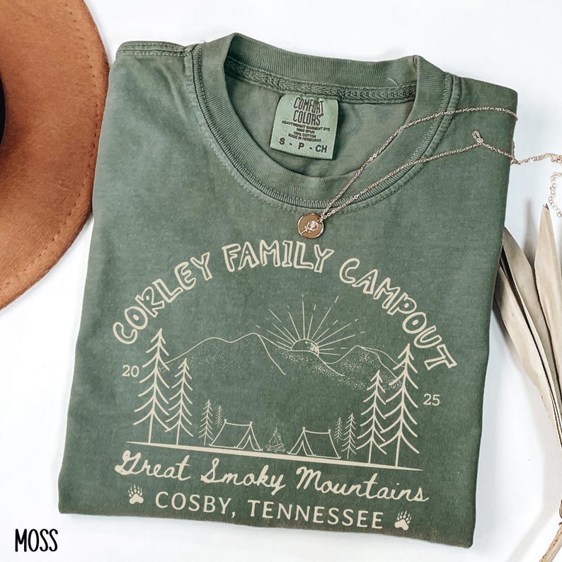 May include: A moss green t-shirt with the text "CORLEY FAMILY CAMPOUT" and "Great Smoky Mountains, Cosby, Tennessee" printed on it. The design includes trees, mountains, and tents. A silver necklace with a pendant is on the shirt.