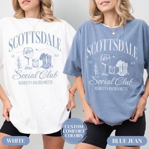May include: Two women wearing white and blue jean t-shirts with a graphic design that says "Scottsdale Social Club" with a cactus, cowboy boots, and a glass of wine. The text also says "Est. 2024" and "Madelyn's Bachelorette".