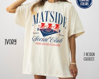 Op de afbeelding: Een wit T-shirt met een grafisch ontwerp dat "Matside Social Club" zegt in een rood-blauw kleurenschema. De grafiek toont drie tuinstoelen en een voetbalveld. De tekst "Fairview" en "Talcons" bevindt zich aan beide kanten van de grafiek. De tekst "Where Loud Mouth Fans Park" bevindt zich onder de grafiek.