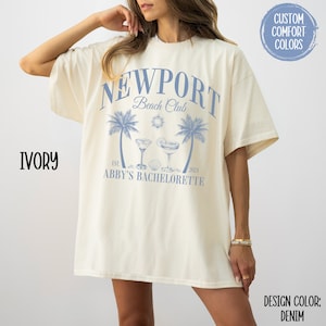 Pu&ograve; includere: Una maglietta bianca oversize con un design grafico blu che dice "Newport Beach Club Est. 2021 Abby's Bachelorette". La grafica include anche palme, un sole e due cocktail.