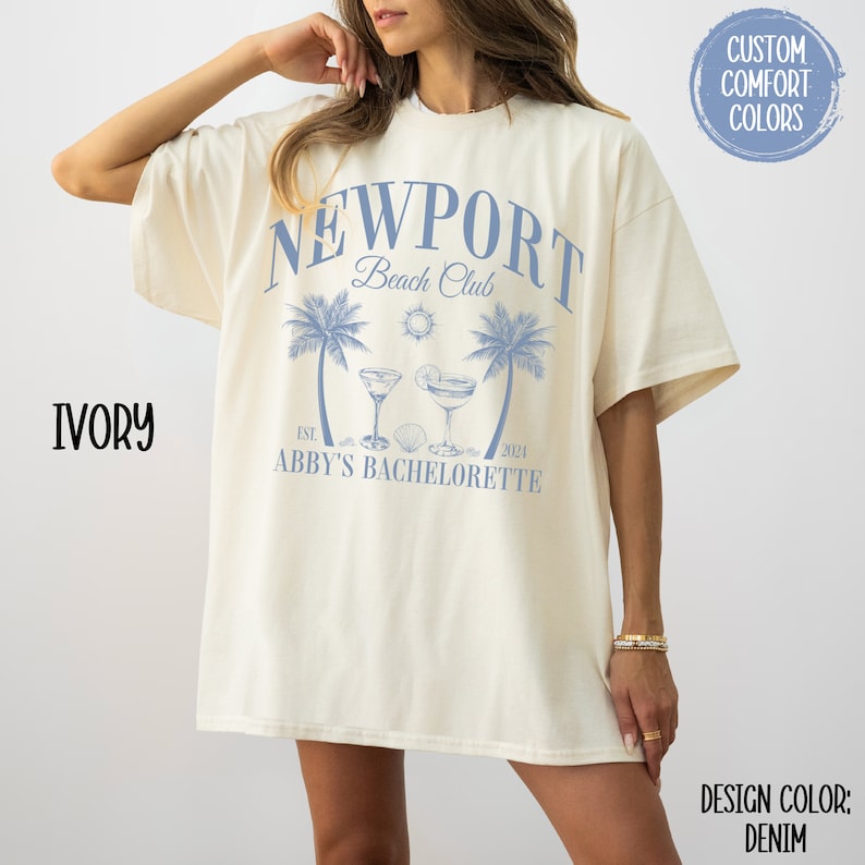 Pu&ograve; includere: Una maglietta bianca oversize con un design grafico blu che dice "Newport Beach Club Est. 2021 Abby's Bachelorette". La grafica include anche palme, un sole e due cocktail.