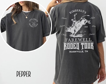 May include: A dark gray t-shirt with a circular graphic on the front that says "The Bach Rodeo Tour". The back of the shirt features a bull riding graphic and the text "Kimberly's Farewell Rodeo Tour Nashville, TN".