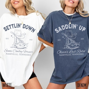 May include: Two oversized t-shirts with a cowboy boot graphic and text. The white t-shirt says "Settlin' Down" and "Same Cowboy Forever" with the year 2025. The blue t-shirt says "Saddlin' Up" and "Olivia's Last Rodeo" with the year 2025.
