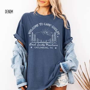 May include: Denim blue t-shirt with a white graphic that reads "WELCOME TO CAMP CORLEY" above a mountain scene with trees and tents. The text "Great Smoky Mountains, Gatlinburg, TN" is below the graphic. The shirt has rolled-up sleeves.