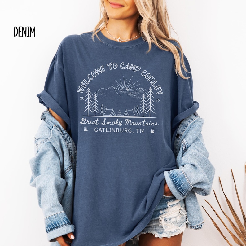 May include: Denim blue t-shirt with a white graphic that reads "WELCOME TO CAMP CORLEY" above a mountain scene with trees and tents. The text "Great Smoky Mountains, Gatlinburg, TN" is below the graphic. The shirt has rolled-up sleeves.