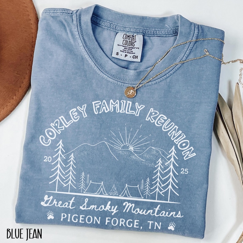 May include: A blue jean Comfort Colors t-shirt with a white graphic that reads "Corley Family Reunion" with a mountain and camping scene. The shirt also has the text "Great Smoky Mountains, Pigeon Forge, TN". A gold necklace with a pendant is on the shirt.