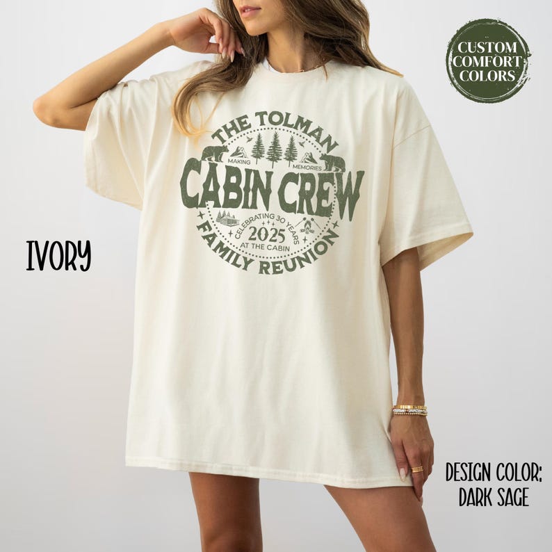 May include: Ivory-colored t-shirt with a dark sage green graphic. The graphic reads "The Tolman Cabin Crew Family Reunion 2025" with bear and tree illustrations. The shirt is oversized and has short sleeves. The design color is dark sage.