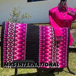 Könnte beinhalten: Ein schwarzer und pinkfarbener Sattelpad mit geometrischem Muster. Der Sattelpad steht auf einem Ständer und der Text "MRS INTERNATIONAL" ist am unteren Rand des Sattelpads sichtbar.