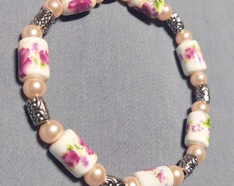 Pode incluir: Um bracelete floral branco e rosa com detalhes prateados. O bracelete é feito de contas de cerâmica com um padrão floral e contas espaçadoras prateadas.