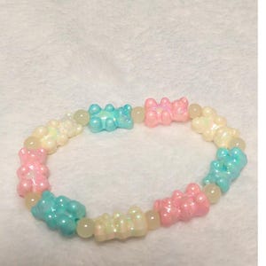 May include: A colorful bracelet featuring gummy bear charms in shades of pink, blue, and yellow. The bracelet is strung with small, round, iridescent beads. The gummy bear charms are evenly spaced around the bracelet.