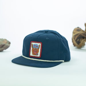 May include: A navy blue flat-brimmed cap with a white rope detail and a rectangular patch. The patch features a cartoon dog wearing a blue hat. The cap is a casual accessory.