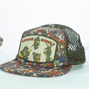 May include: A trucker hat with a brown mesh back and a patterned front featuring leaves in shades of brown, orange, and blue. The hat has a patch with the words "SAVAGE SUPPLY" and cartoon animals playing guitars.