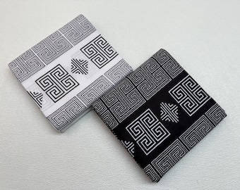 May include: Two folded fabric squares, one white and one black, both featuring a repeating geometric pattern in black and white. The design includes square and rectangular shapes with dotted borders.