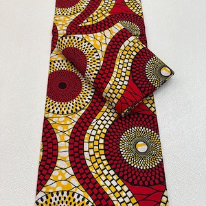 May include: A vibrant fabric with a repeating pattern of red, yellow, and black circular and geometric designs. The fabric is folded over, showcasing the intricate details of the print. The bold colors and patterns suggest an African textile.