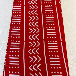 May include: A rectangular piece of red fabric featuring a repeating white pattern of vertical stripes, horizontal dashes, dots, and chevron shapes. The bottom edge includes the printed text 100% COTTON GUARANTEED BLOCK PRINTS SUPER GLOBAL.