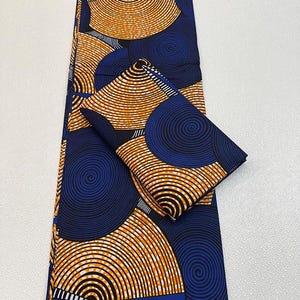 May include: A folded fabric with a repeating pattern of orange and white concentric circles on a navy blue background. A smaller, folded piece of the same fabric is also present. The fabric has a bold, geometric design.
