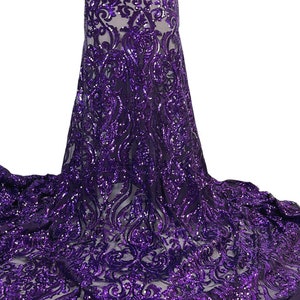May include: A close-up of a flowing fabric with a detailed, ornate pattern. The fabric is a deep purple color, covered in shimmering sequins. The design features swirling, floral-like motifs, creating a luxurious and elegant appearance. The fabric appears to be a high-end textile.
