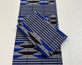 May include: Blue, black, and white patterned fabric, folded and displayed. The fabric features a repeating geometric design with horizontal stripes and diamond-like shapes. The folded piece sits atop a longer, unfolded section.
