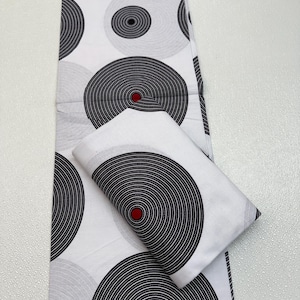May include: White fabric with a repeating pattern of black and gray concentric circles, each with a red center. A folded piece of the same fabric is also present, showcasing the design.