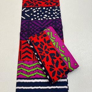 May include: A vibrant textile with bold geometric patterns in red, purple, navy, and green. The fabric features a mix of shapes, including dots, stripes, and abstract designs. Two folded pieces of the fabric are on top of the larger piece.