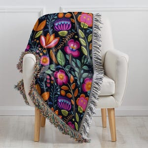 May include: A black woven throw blanket with a colorful floral pattern. The blanket has a fringed edge and is draped over a white armchair.