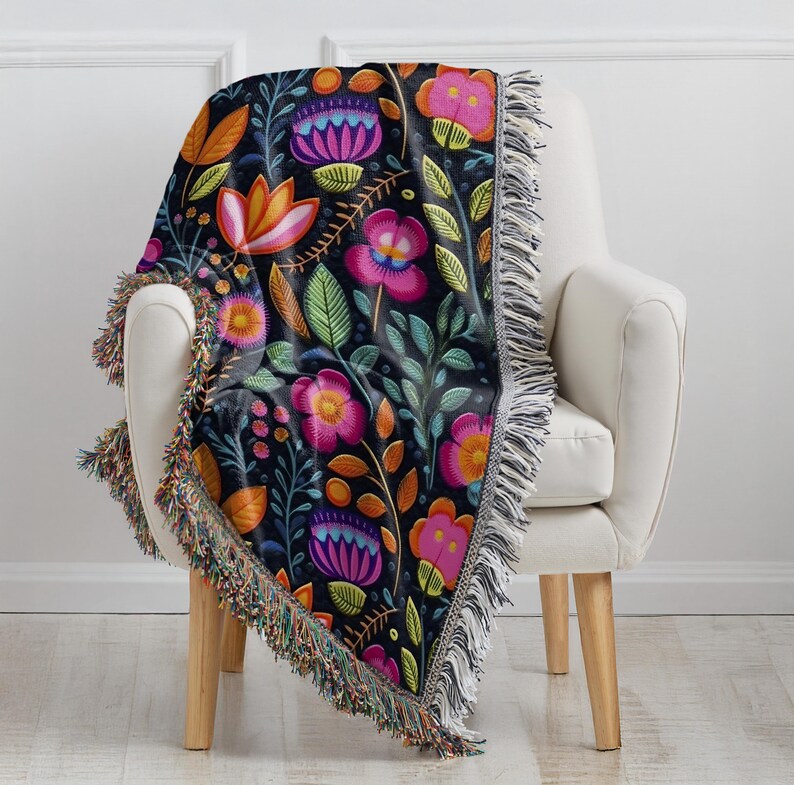 May include: A black woven throw blanket with a colorful floral pattern. The blanket has a fringed edge and is draped over a white armchair.